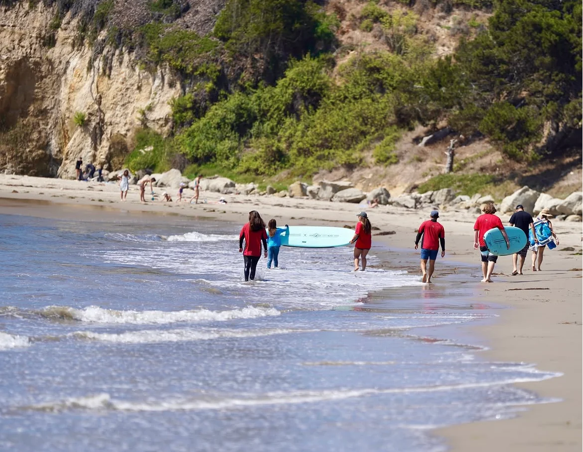 Surfers and volunteers carrying boards along the water beneath coastal cliffs