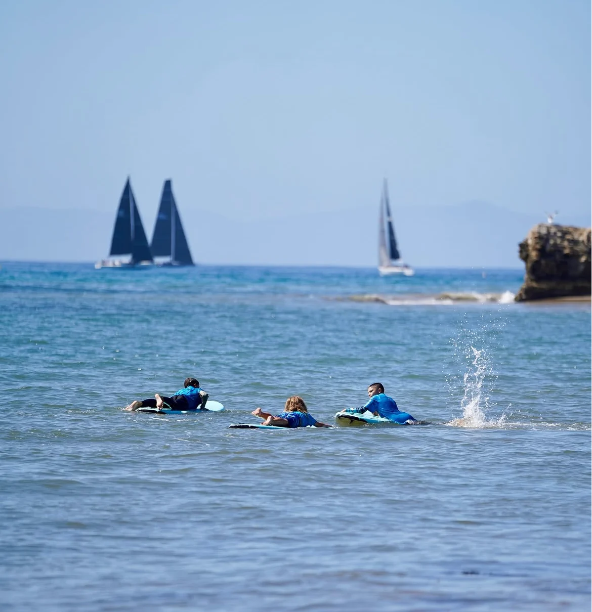 Three surfers paddling toward sailboats along the coastline