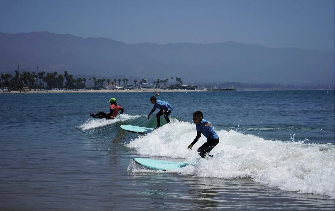Surfers sharing a wave with mountains and palms along the coast behind them